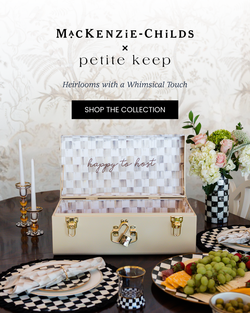 Heirlooms with a Whimsical Touch. Mackenzie-Childs x Petite Keep collection with a heirloom beige trunk on a table setting with Mocha Check print with Happy to Host embroidered on it. Shop the collection.